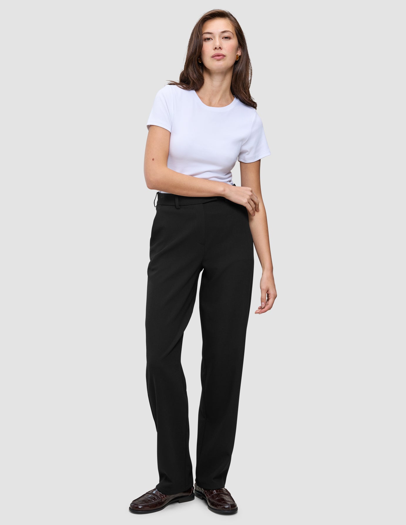 Eine Frau in einem weißen T-Shirt mit Rundhalsausschnitt, einer schwarzen Women GO2-Hose in gerader Passform und dunkelbraunen Loafern, die ein minimalistisches skandinavisches Design widerspiegeln.